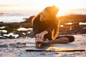 A woman doing yoga on the beach at sunset; rocks and the ocean visible in the background