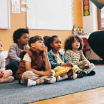 Elementary school students sit on a carpet and listen to a story from their teacher.