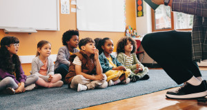 Elementary school students sit on a carpet and listen to a story from their teacher. 
