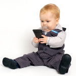 A baby wearing a suit with a "binkie" in his mouth, hoding and staring at a cellphone