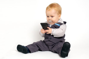 A baby wearing a suit with a "binkie" in his mouth, hoding and staring at a cellphone