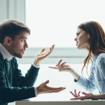 A man and woman sit across a small table talking with each other. He shrugs his shoulders in puzzlement, she points in irritation.