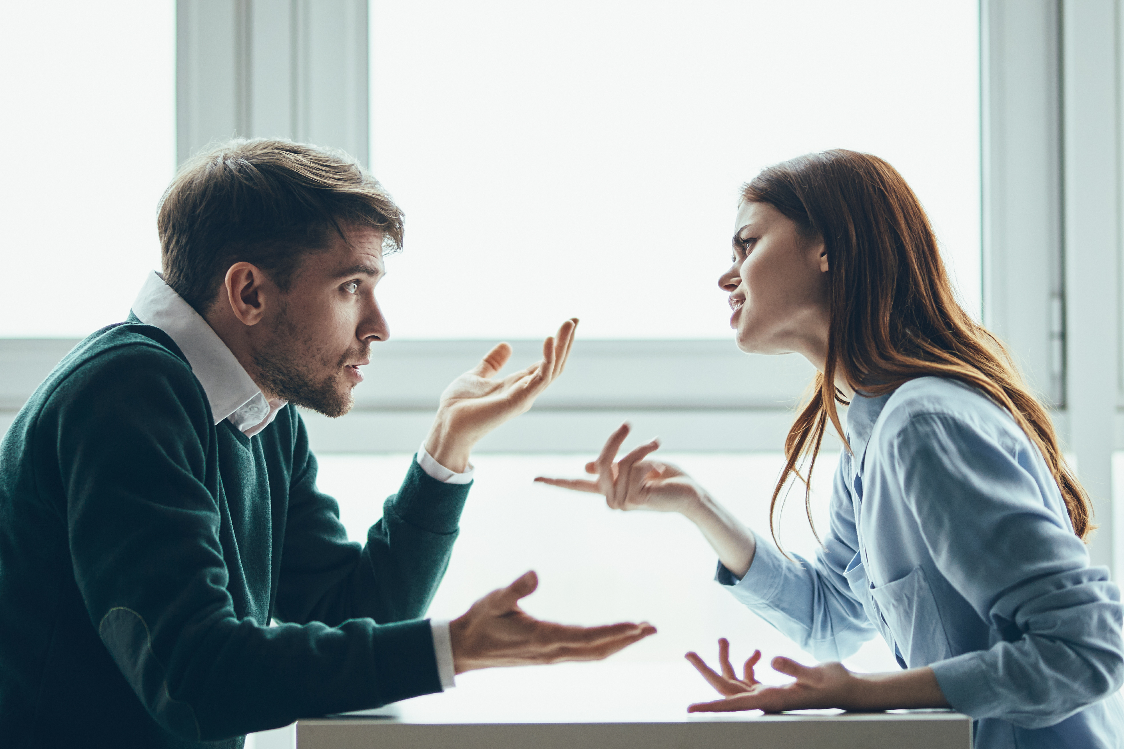 A man and woman sit across a small table talking with each other. He shrugs his shoulders in puzzlement, she points in irritation.