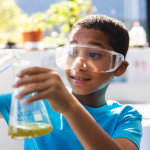 A young student wearing plastic goggles carefully pours something into a beaker slightly filled with green liquid