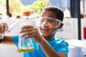 A young student wearing plastic goggles carefully pours something into a beaker slightly filled with green liquid