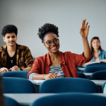 A college age student smiling and raising her hand to ask a question.