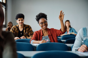 A college age student smiling and raising her hand to ask a question.