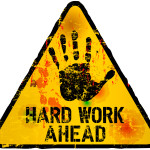 A triangular yellow road sign reading "Hard Work Ahead." the sign has a large hand print on it, and smudges of dirt or grime.